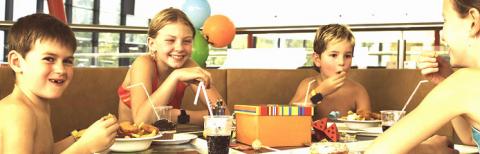 Vier Kinder in Badeanzügen sitzen an einem Tisch, essen und trinken aus Strohhalmen, im Hintergrund sind Luftballons und eine Geschenkbox zu sehen.