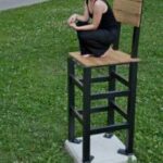 Eine Frau hockt barfuß auf einem übergroßen Stuhl aus Holz und Metall, der auf einem Betonsockel im Freien auf einer Wiese steht.