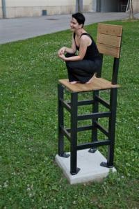 Eine Frau hockt barfuß auf einem übergroßen Stuhl aus Holz und Metall, der auf einem Betonsockel im Freien auf einer Wiese steht.