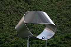 Eine große metallene Möbiusband-Skulptur steht im Freien vor einem grünen, begrünten Hintergrund.