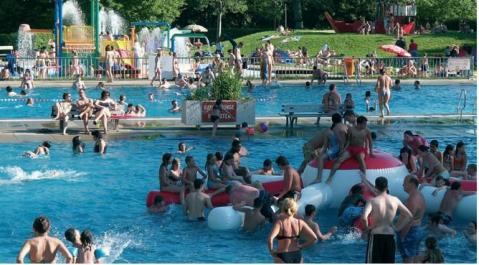 Ein überfülltes öffentliches Freibad mit Menschen, die bei sonnigem Wetter schwimmen, faulenzen und auf einer großen aufblasbaren Struktur spielen.