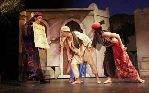 Vier Schauspieler in farbenfrohen, historischen Kostümen spielen auf der Bühne eine dramatische Szene, wobei einer eine Schriftrolle hält und die anderen vor einer gemalten Gebäudekulisse lehnen.