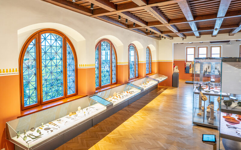 Ausstellungsraum des Museums mit großen Bogenfenstern, Holzfußboden und Vitrinen mit verschiedenen Artefakten an den Wänden und in der Mitte.
