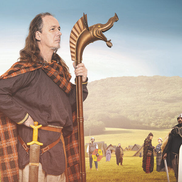 Ein Mann in historischer Kleidung und mit einem Stab in der Hand steht auf einem Feld mit anderen Personen in ähnlicher Kleidung und mit Schilden, mit Zelten und Hügeln im Hintergrund.