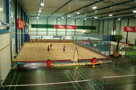 Sporthalle mit einem Sandvolleyballfeld, das von einem Zaun umgeben ist, und weiteren Feldern und Geräten im Vordergrund.