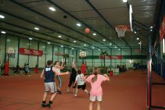 Eine Gruppe von Kindern spielt drinnen auf einem Platz Basketball. Ein Kind wirft den Ball in Richtung des Korbes, während die anderen zuschauen oder versuchen, ihn zu blocken.