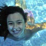 Ein Kind mit langen dunklen Haaren schwimmt in einem Pool unter Wasser und lächelt in die Kamera, während das Sonnenlicht Muster auf dem Poolboden erzeugt.