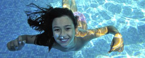 Ein Kind mit langen dunklen Haaren schwimmt in einem Pool unter Wasser und lächelt in die Kamera, während das Sonnenlicht Muster auf dem Poolboden erzeugt.