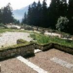 Ein Hügelfriedhof mit steinernen Grabsteinen, umgeben von Grün und Bäumen, mit den Bergen im Hintergrund.