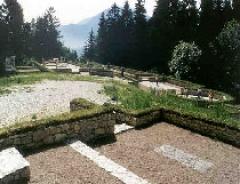 Ein Hügelfriedhof mit steinernen Grabsteinen, umgeben von Grün und Bäumen, mit den Bergen im Hintergrund.