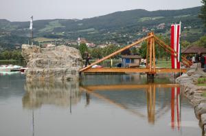 Eine Holzbrücke überquert ein ruhiges Gewässer mit einer felsigen Struktur und einer großen rot-weiß senkrecht gestreiften Flagge in der Nähe; im Hintergrund sind Hügel und Häuser zu sehen.