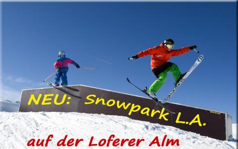 Zwei Skifahrer in farbenfroher Ausrüstung springen von einer Rampe im Snowpark L.A., Loferer Alm, unter blauem Himmel. Ein Text in deutscher Sprache wirbt für den neuen Standort des Snowparks.