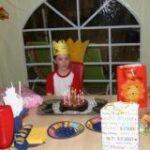 Ein Kind, das eine Krone trägt, sitzt an einem Tisch mit Geburtstagskuchen, Geschenken und anderen Kindern in einem geschmückten Raum.