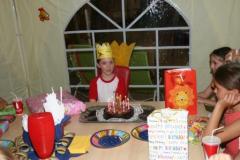 Ein Kind, das eine Krone trägt, sitzt an einem Tisch mit Geburtstagskuchen, Geschenken und anderen Kindern in einem geschmückten Raum.