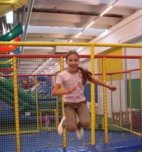 Ein junges Mädchen springt auf einem Trampolin in einem Indoor-Spielbereich mit bunten Kletterstrukturen und Sicherheitsnetzen.