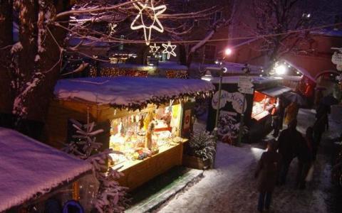 Die verschneiten Marktstände unter freiem Himmel sind nachts mit Lichterketten und Sternen beleuchtet, und zwischen den Ständen laufen ein paar Leute auf einem verschneiten Weg.