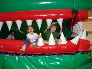 Zwei Kinder sitzen und spielen auf einer aufblasbaren Struktur, die wie das offene Maul eines großen grünen Wesens mit weißen spitzen Zähnen aussieht.