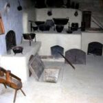 Historische Küche mit weiß getünchten Wänden, alten Kochtöpfen und Öfen; ein Holzschemel und eine offene Kellerluke auf dem Steinboden sind zu sehen.