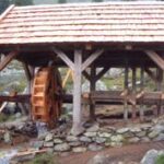 Eine hölzerne Wassermühle mit einem Schindeldach steht auf einem steinernen Fundament in einer ländlichen Umgebung mit Bäumen im Hintergrund.