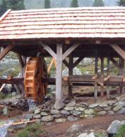Eine hölzerne Wassermühle mit einem Schindeldach steht auf einem steinernen Fundament in einer ländlichen Umgebung mit Bäumen im Hintergrund.