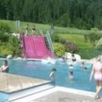 Kinder spielen und schwimmen in einem Außenpool mit einer rosafarbenen Doppelrutsche, umgeben von Grün und Bäumen im Hintergrund.