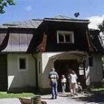 Eine kleine Gruppe von Menschen steht an einem sonnigen Tag vor einem malerischen Gebäude im Landhausstil, umgeben von Bäumen und Grünflächen.