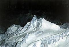 Ein schneebedeckter Berggipfel hebt sich scharf von einem dunklen Hintergrund ab, umgeben von zerklüftetem, eisigem Terrain.