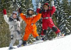 Drei Kinder in bunten Schneeanzügen stehen mit erhobenen Armen auf einem verschneiten Hang, umgeben von schneebedeckten Bäumen.
