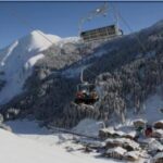 Skifahrer fahren mit einem Sessellift über ein verschneites Bergdorf, mit schneebedeckten Bäumen und Bergen im Hintergrund.