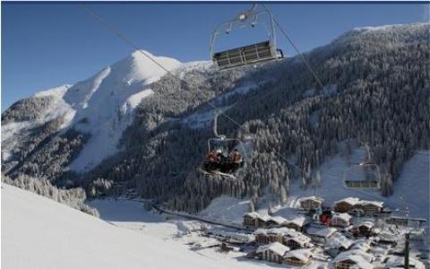 Skifahrer fahren mit einem Sessellift über ein verschneites Bergdorf, mit schneebedeckten Bäumen und Bergen im Hintergrund.