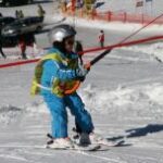 Ein Kind in blauer Skibekleidung und mit Helm benutzt auf einer verschneiten Piste einen Schlepplift und hält sich am Seil fest, während es auf Skiern steht.