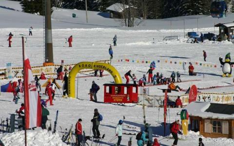 Kinder und Erwachsene fahren Ski und spielen in einem verschneiten Gebiet mit farbenfrohen Dekorationen, einer roten Zugstruktur und verschiedenen Schildern. Im Hintergrund sind schneebedeckte Bäume zu sehen.