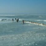 Holzpfähle ragen in ein gefrorenes Gewässer unter einem strahlend blauen Himmel, auf dem Eisflächen und sanfte Wellen zu sehen sind.