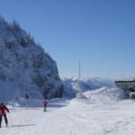 Zwei Personen fahren auf einer verschneiten Bergpiste unter strahlend blauem Himmel Ski, im Hintergrund sind schneebedeckte Bäume und Gebäude zu sehen.