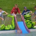 Vier Jungen spielen an einer kleinen Rutsche mit großen grünen Froschfiguren neben einem flachen Wasserbecken.