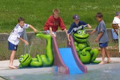 Vier Jungen spielen an einer kleinen Rutsche mit großen grünen Froschfiguren neben einem flachen Wasserbecken.