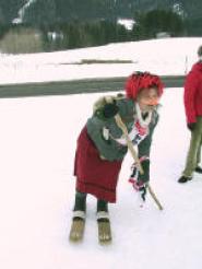 Eine Person in traditioneller Kleidung mit Kopftuch und Holzschuhen steht mit einem Stock in der Hand im Schnee, eine weitere Person ist teilweise im Hintergrund zu sehen.