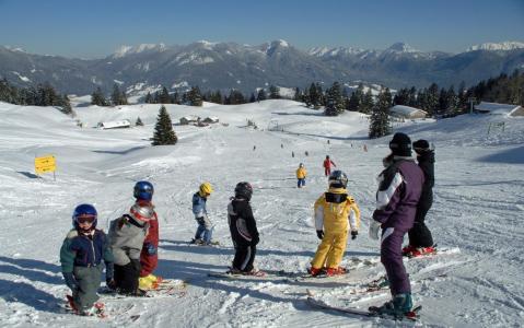 Eine Gruppe von Kindern in Winterkleidung steht auf einer verschneiten Skipiste mit Bergen und Bäumen im Hintergrund.