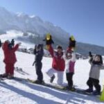 Sechs Kinder in Winterkleidung stehen auf Snowboards in einer Reihe auf einem verschneiten Hang und heben ihre Arme, mit Bergen und blauem Himmel im Hintergrund.