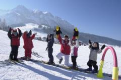 Sechs Kinder in Winterkleidung stehen auf Snowboards in einer Reihe auf einem verschneiten Hang und heben ihre Arme, mit Bergen und blauem Himmel im Hintergrund.