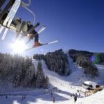 An einem sonnigen Tag fahren Skifahrer mit einem Sessellift über einen verschneiten Berghang, unter dem Bäume, Skifahrer und eine Skihütte zu sehen sind.