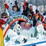 Eine Gruppe von Kindern in Winterkleidung fährt an einem verschneiten Tag lächelnd und winkend mit einem bunten Zug mit der Aufschrift "Rote Teufel Express".