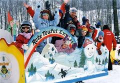 Eine Gruppe von Kindern in Winterkleidung fährt an einem verschneiten Tag lächelnd und winkend mit einem bunten Zug mit der Aufschrift "Rote Teufel Express".