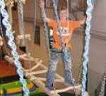 Ein Kind, das einen Sicherheitsgurt trägt, läuft auf einer Hängeseilbrücke in einem Indoor-Abenteuerspielplatz.