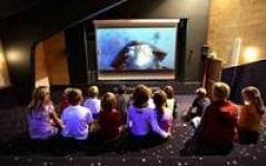 Eine Gruppe von Kindern sitzt in einem schwach beleuchteten Raum auf dem Boden und sieht sich einen auf eine große Leinwand projizierten Film an.