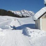 Eine kleine schneebedeckte Kapelle steht neben einem verschneiten Weg, auf dem Menschen Ski fahren oder rodeln, umgeben von Bäumen und Bergen im Hintergrund unter einem klaren blauen Himmel.