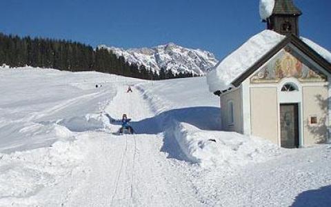 Eine kleine schneebedeckte Kapelle steht neben einem verschneiten Weg, auf dem Menschen Ski fahren oder rodeln, umgeben von Bäumen und Bergen im Hintergrund unter einem klaren blauen Himmel.