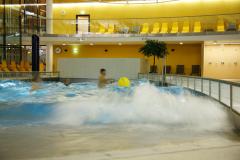 Mehrere Personen spielen in einem Hallenbad mit künstlichen Wellen; im Hintergrund sind gelbe Wände und leere Liegestühle zu sehen.