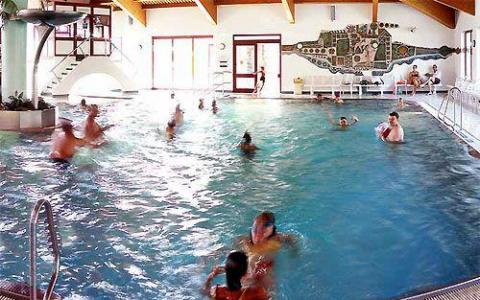 Menschen schwimmen und spielen in einem Hallenbad mit natürlichem Licht, das durch große Fenster einfällt; an der Wand im Hintergrund ist ein Wandgemälde zu sehen.