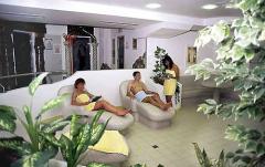 Drei Personen in Handtüchern entspannen sich auf Liegestühlen in einem Spa-ähnlichen Raum mit Pflanzen und gefliesten Böden.